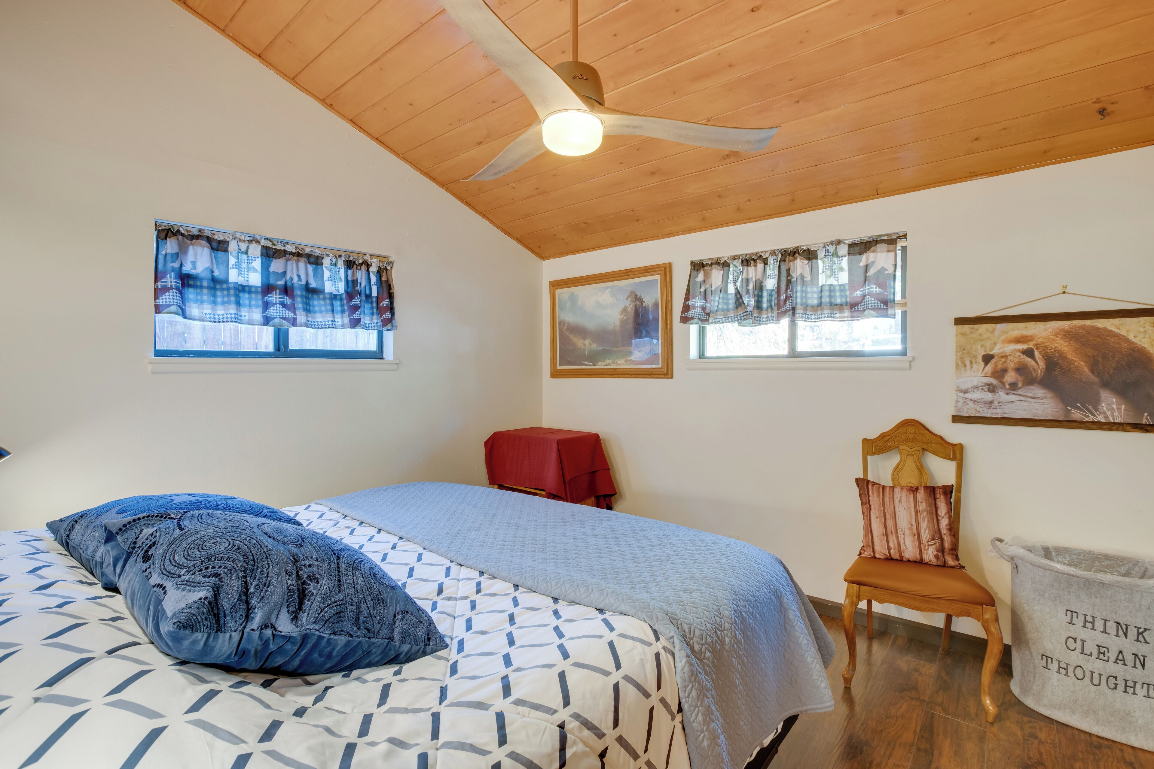 A bedroom with a bed, a wooden ceiling fan, a red chair, and a wall-mounted trash bin.