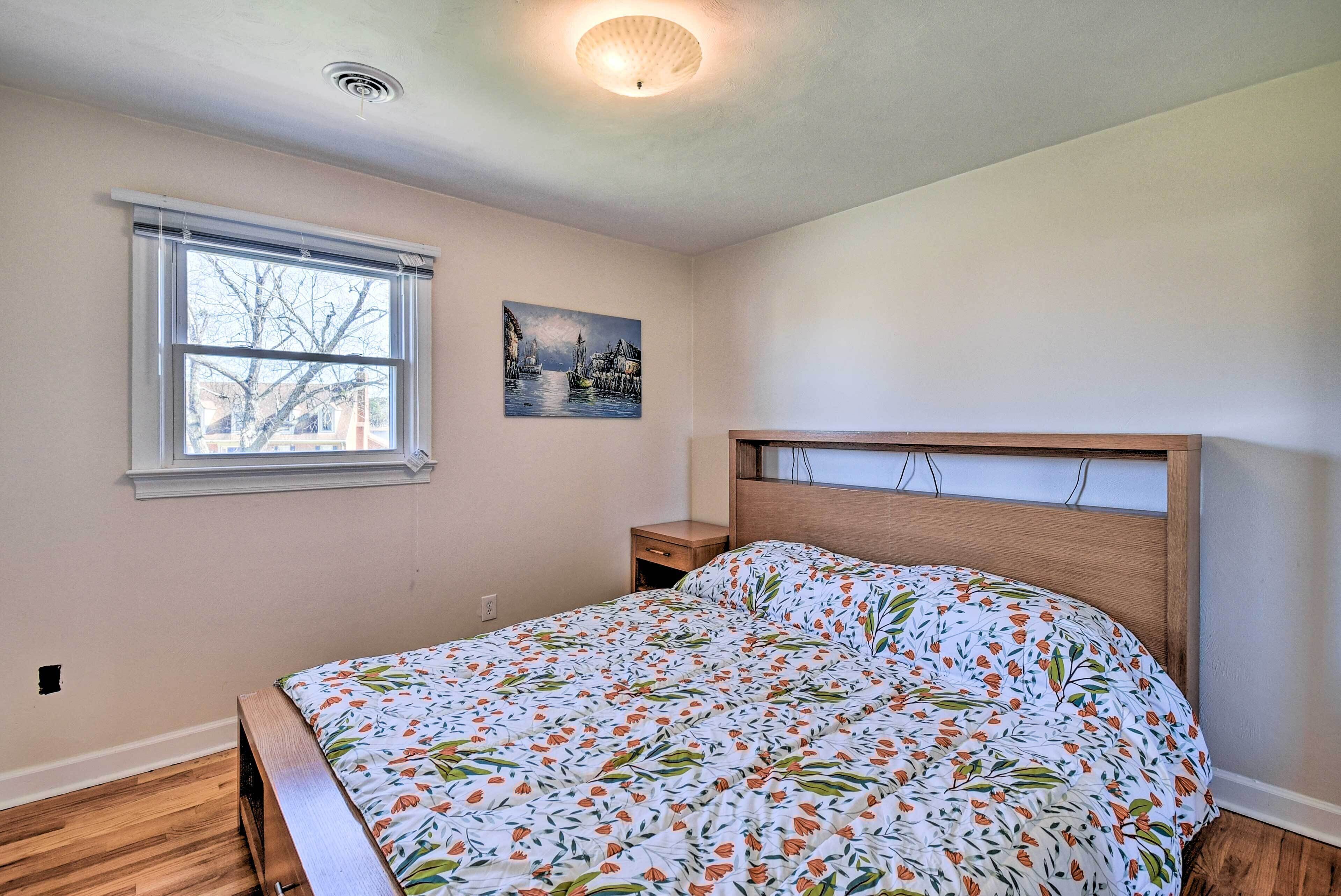 A bedroom with a wooden headboard, a bed with a floral bedspread, a nightstand, a window with blinds, and a wall-mounted painting.