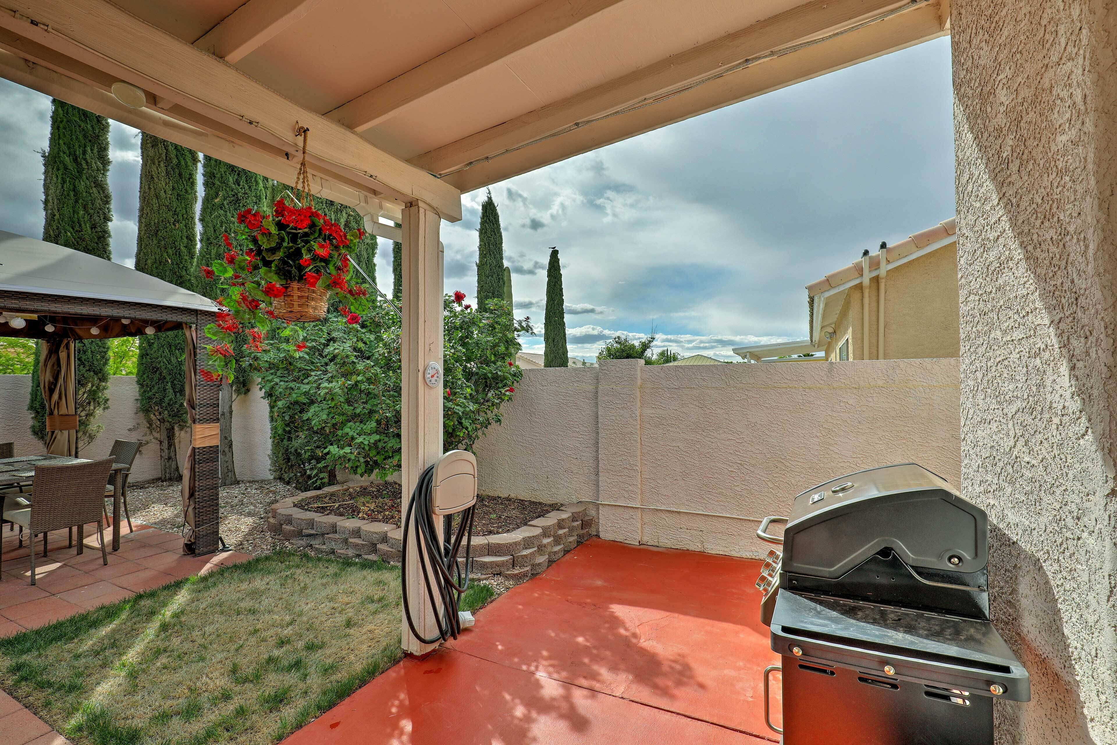 A covered patio with a grill, a dining table, and hanging red flowers.