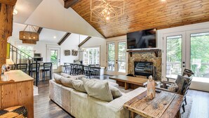 A spacious living area with a wooden ceiling, a stone fireplace, and a large window overlooking greenery. - The Ultimate Luxury Retreat on Lake Burton! (Clayton)
