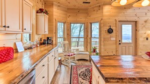 Un intérieur de chalet chaleureux avec des murs en bois, un coin cuisine doté d’un fauteuil à bascule et de grandes fenêtres offrant une vue sur l’extérieur.