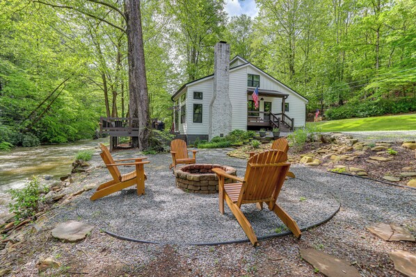 Un chalet doté d’une cheminée en pierre, entouré de fauteuils Adirondack, situé dans une zone boisée avec une rivière en arrière-plan.