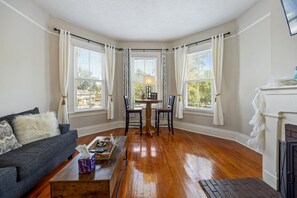 A cozy living room with a sofa, a coffee table, and a fireplace. There are two windows with curtains, a lamp on the table, and a view of the outside.