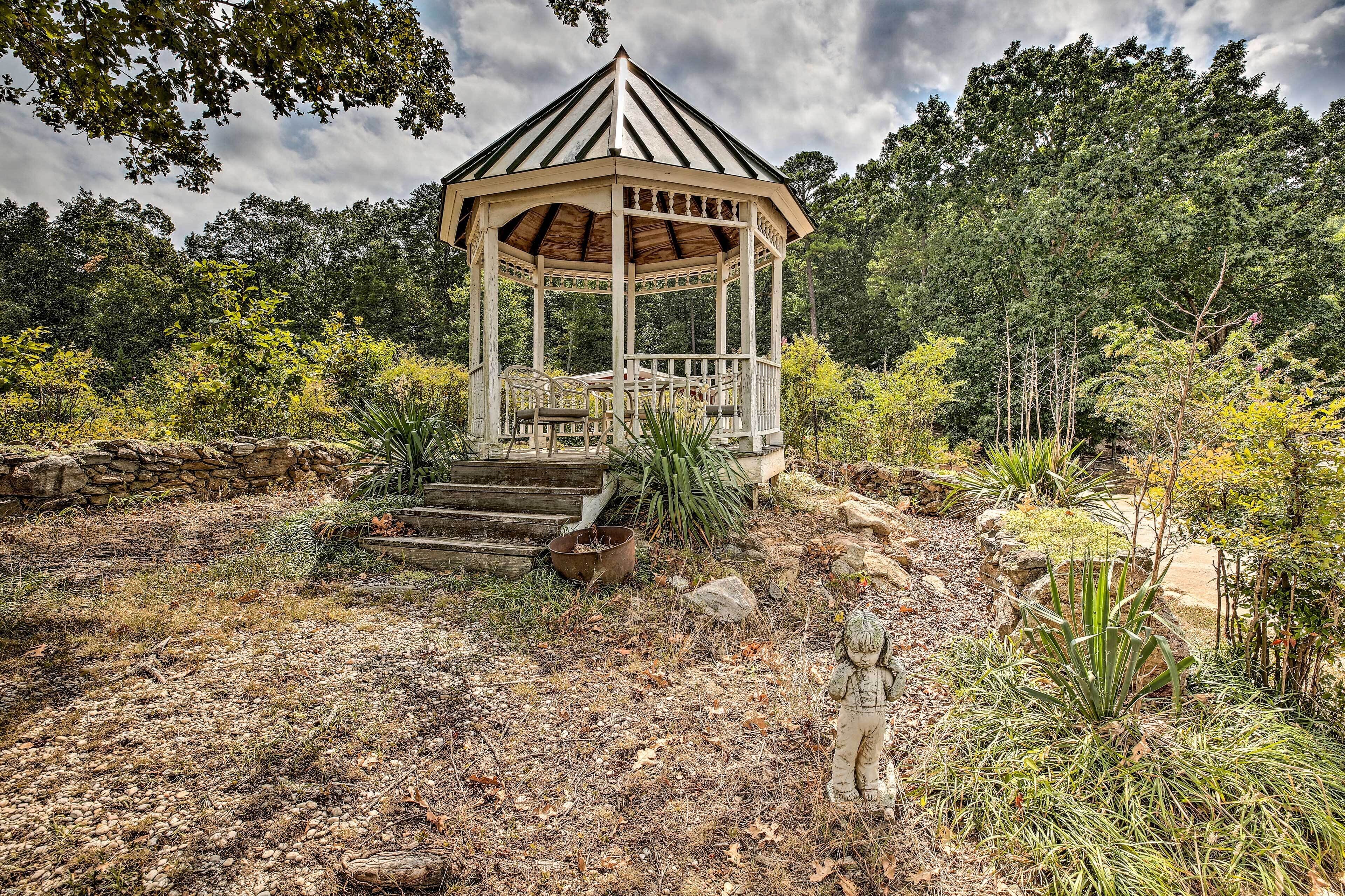 A gazebo with a wooden roof and white walls, surrounded by a garden with various plants and a stone wall.