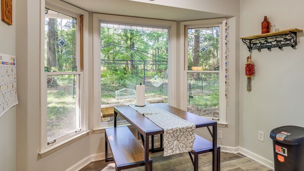 A room with a wooden table and benches, large windows with a view of trees, a calendar on the wall, and a shelf with decorative items.