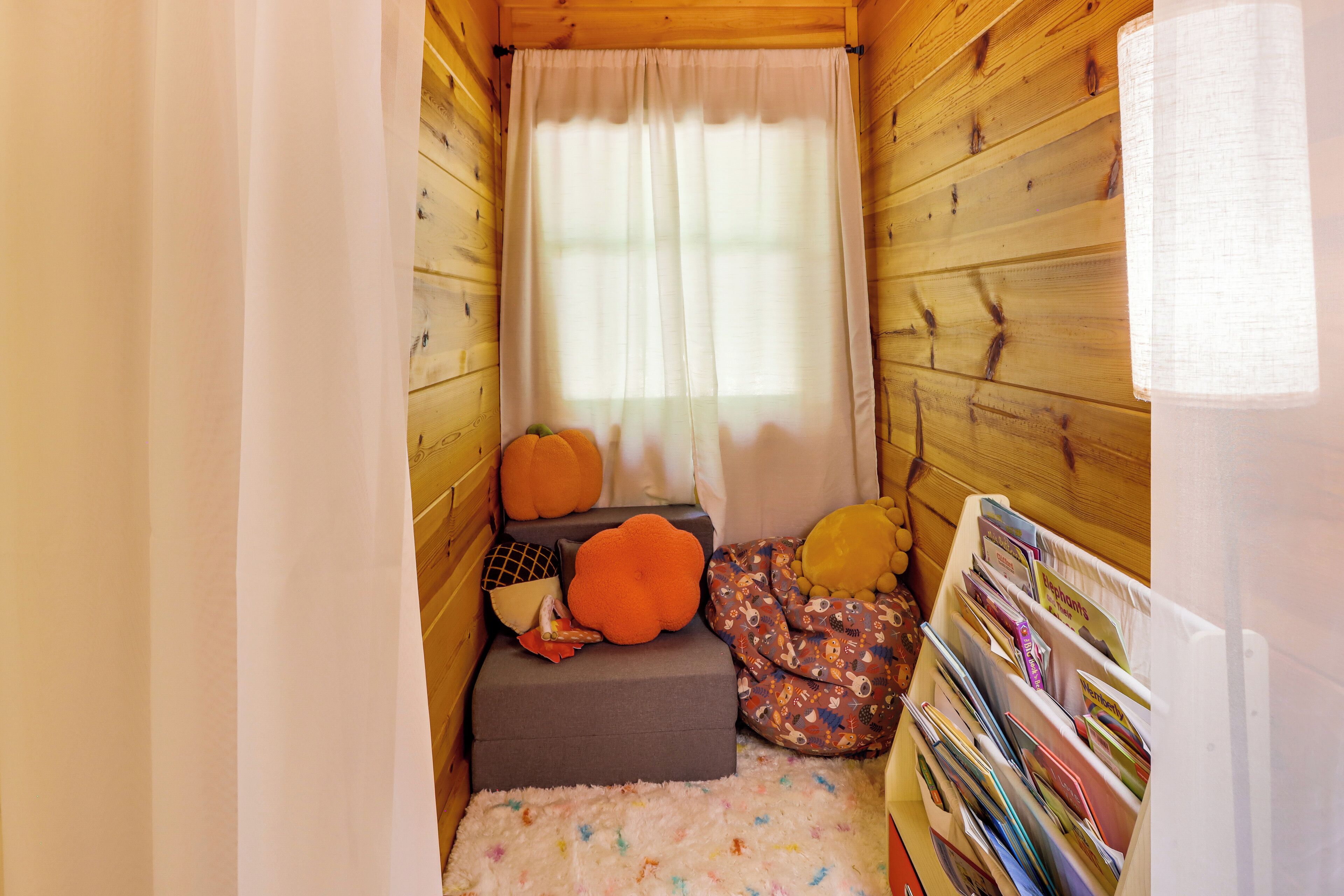 Une pièce chaleureuse aux murs en bois, une fenêtre ornée de rideaux blancs, un petit coin salon avec des coussins et une bibliothèque remplie de livres.
