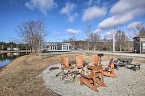 A gravel patio with Adirondack chairs and a fire pit, a body of water, and a building with a green roof in the background. - Peaceful Deck & Pool Access! Blue Ridge Tiny Home (Morganton)