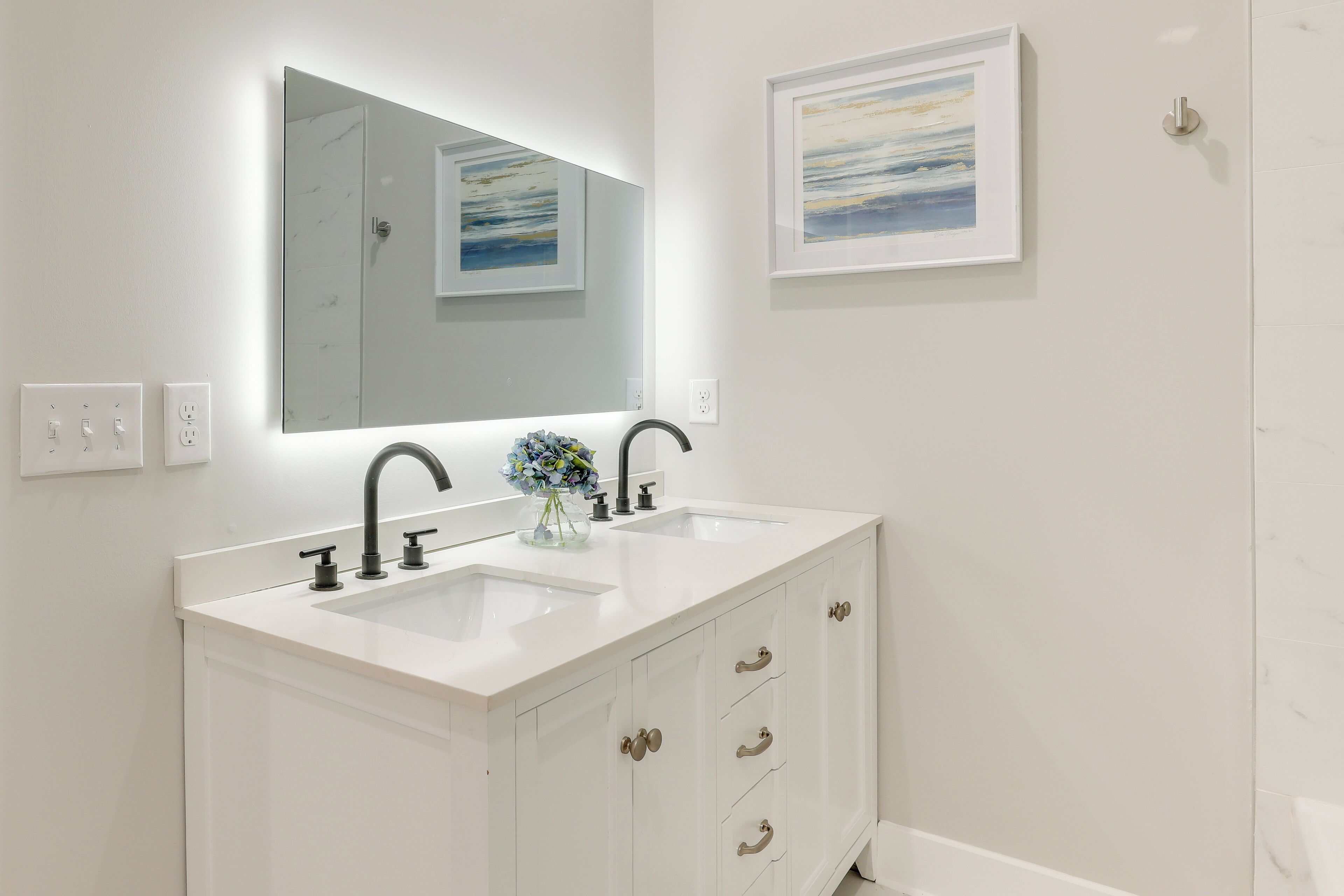A bathroom with a white vanity, two sinks, a large mirror, and framed artwork on the wall.