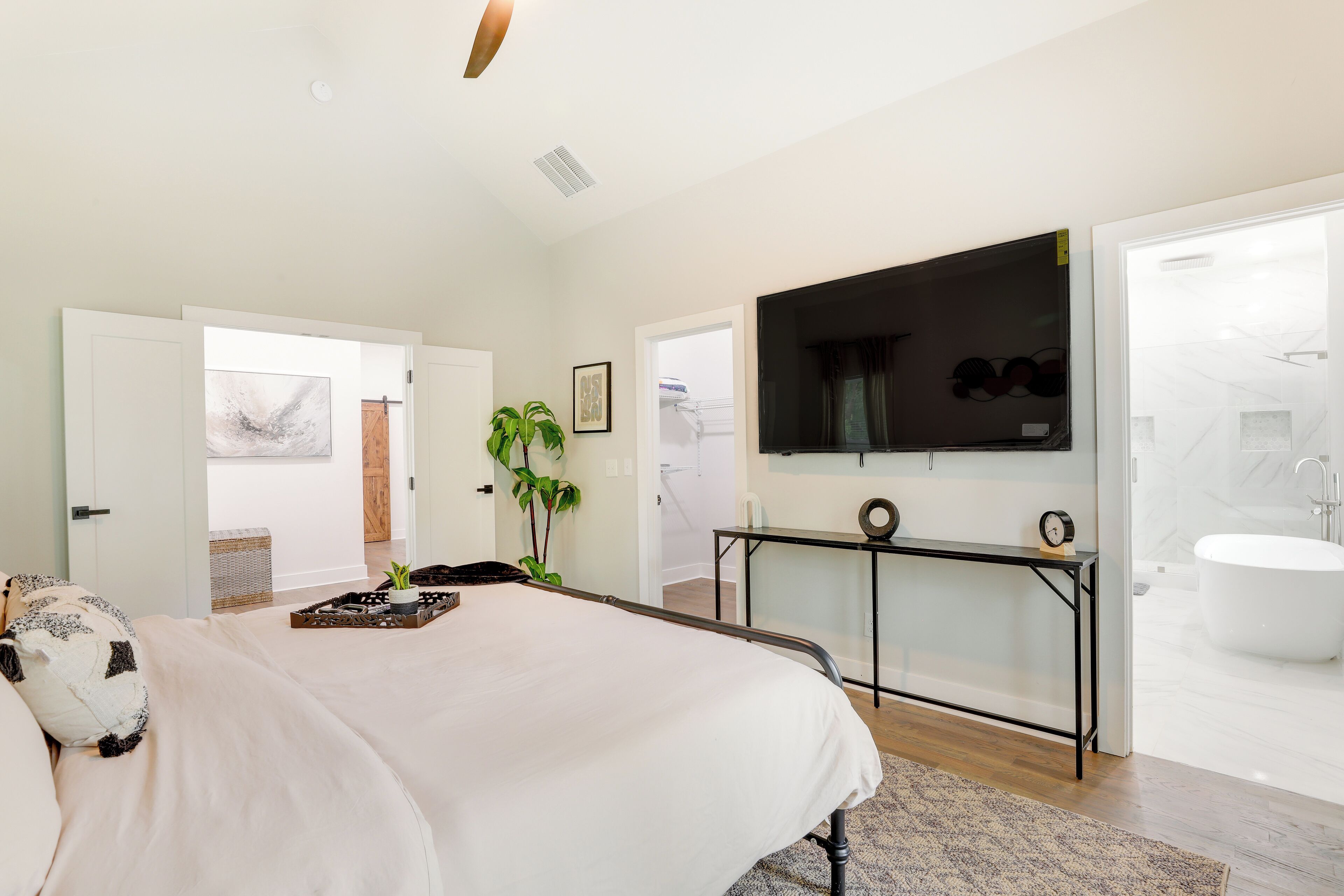 A bedroom with a large bed, a TV mounted on the wall, a bedside table, and a bathroom with a bathtub and toilet visible through an open door.