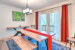 Uma área de refeições com mesa de madeira e bancos, um candelabro e uma janela grande com cortinas vermelhas.