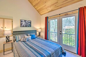 Quarto com cama grande, duas mesas de cabeceira com abajures, teto de madeira, janela com cortinas e vista para árvores.
