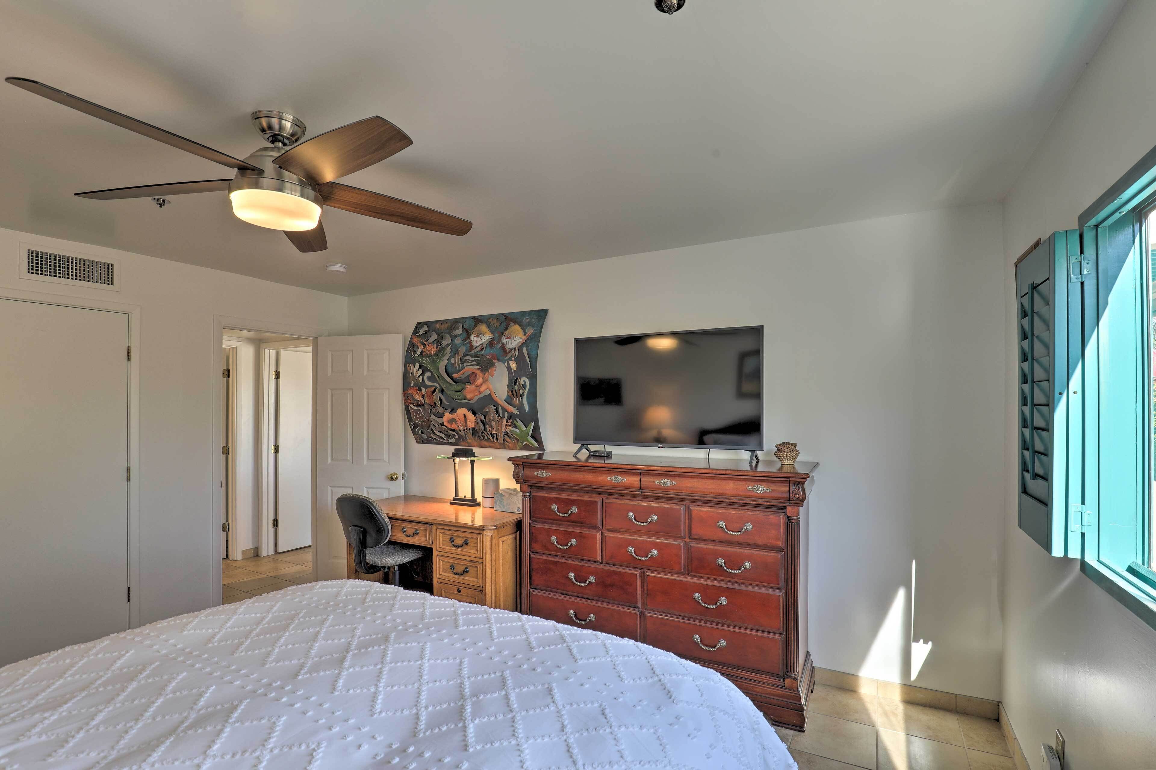 Une chambre à coucher comprenant un lit, un ventilateur de plafond, une commode, un bureau avec une chaise, un téléviseur et une fenêtre avec des stores.