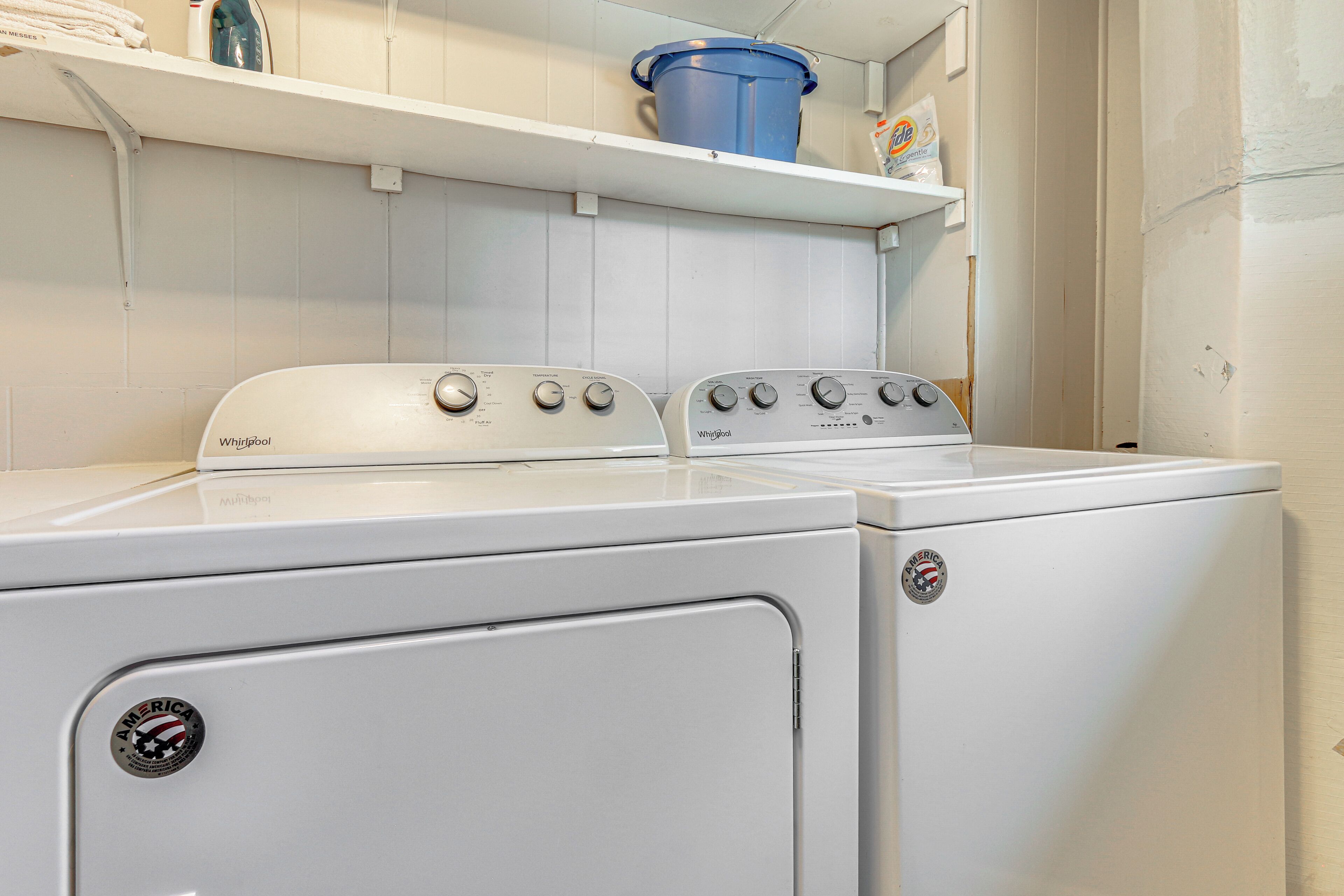 Une buanderie équipée de deux lave-linge Whirlpool blancs et d’un seau bleu posé sur une étagère.