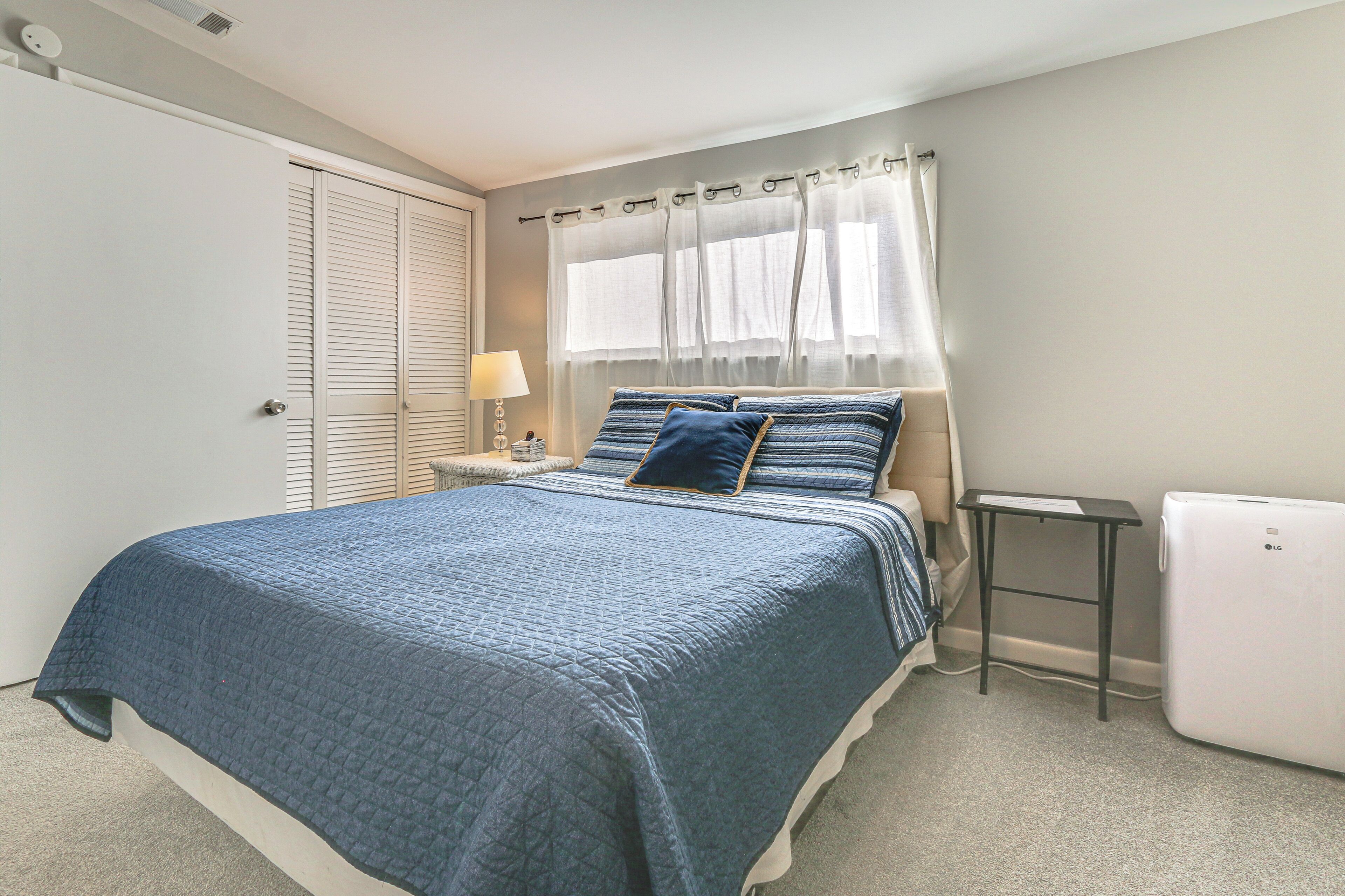 A bedroom with a bed, a nightstand, a lamp, a window with curtains, and a white appliance.