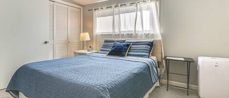 A bedroom with a bed, a nightstand, a lamp, a window with curtains, and a white appliance.