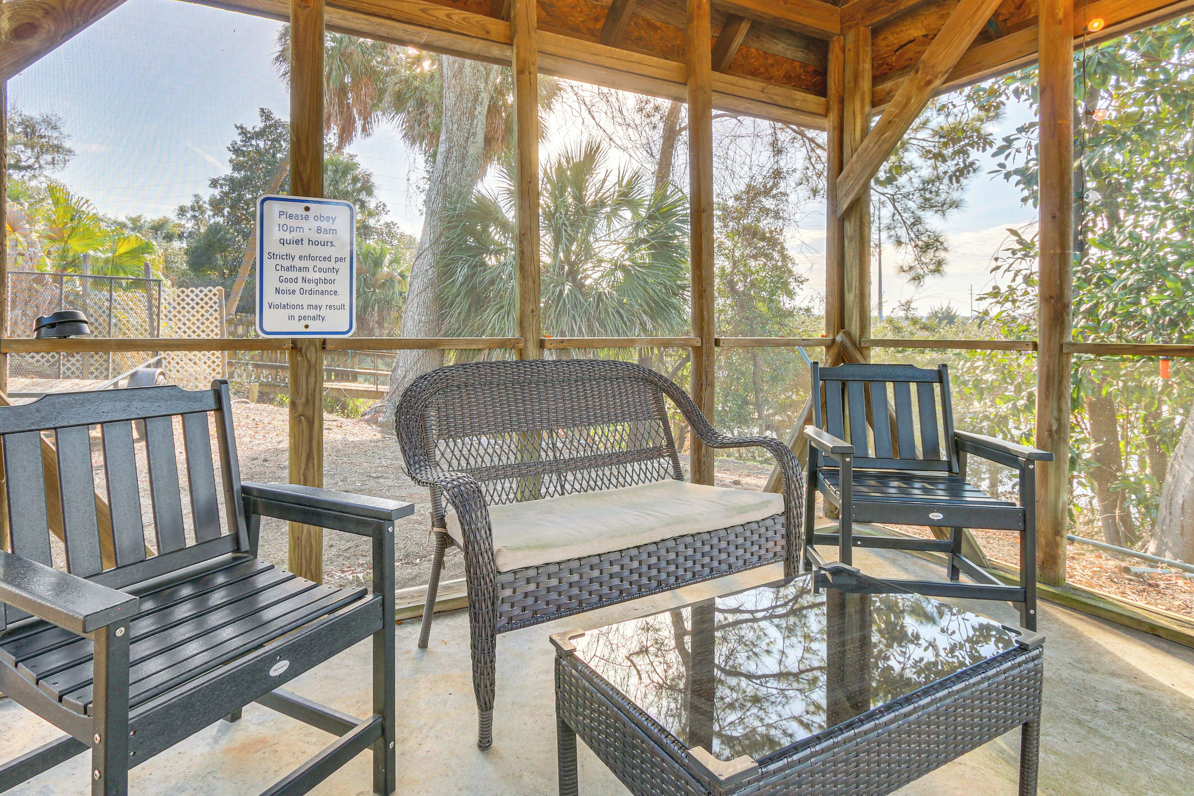 A covered outdoor seating area with wicker furniture and a glass table, surrounded by a wooden fence and palm trees.