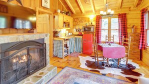 A rustic cabin interior with a stone fireplace, wooden walls, a kitchen area, and a dining table set for four. - 'little Fox Den Ellijay Cabin Rental w/ Hot Tub! (Ellijay)
