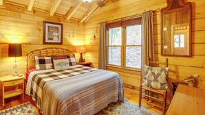 A wooden cabin bedroom with a bed, nightstands, a chair, and a window with curtains. - 'little Fox Den Ellijay Cabin Rental w/ Hot Tub! (Ellijay)