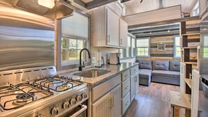 A modern kitchen with a gas stove, stainless steel appliances, and a built-in oven. There is a gray sofa in the adjacent living area, and the space features wooden beams and large windows. - Blairsville Tiny Home w/ Covered Furnished Deck! (Blairsville)