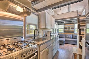 A modern kitchen with a gas stove, stainless steel appliances, and a built-in oven. There is a gray sofa in the adjacent living area, and the space features wooden beams and large windows. - Blairsville Tiny Home w/ Covered Furnished Deck! (Blairsville)