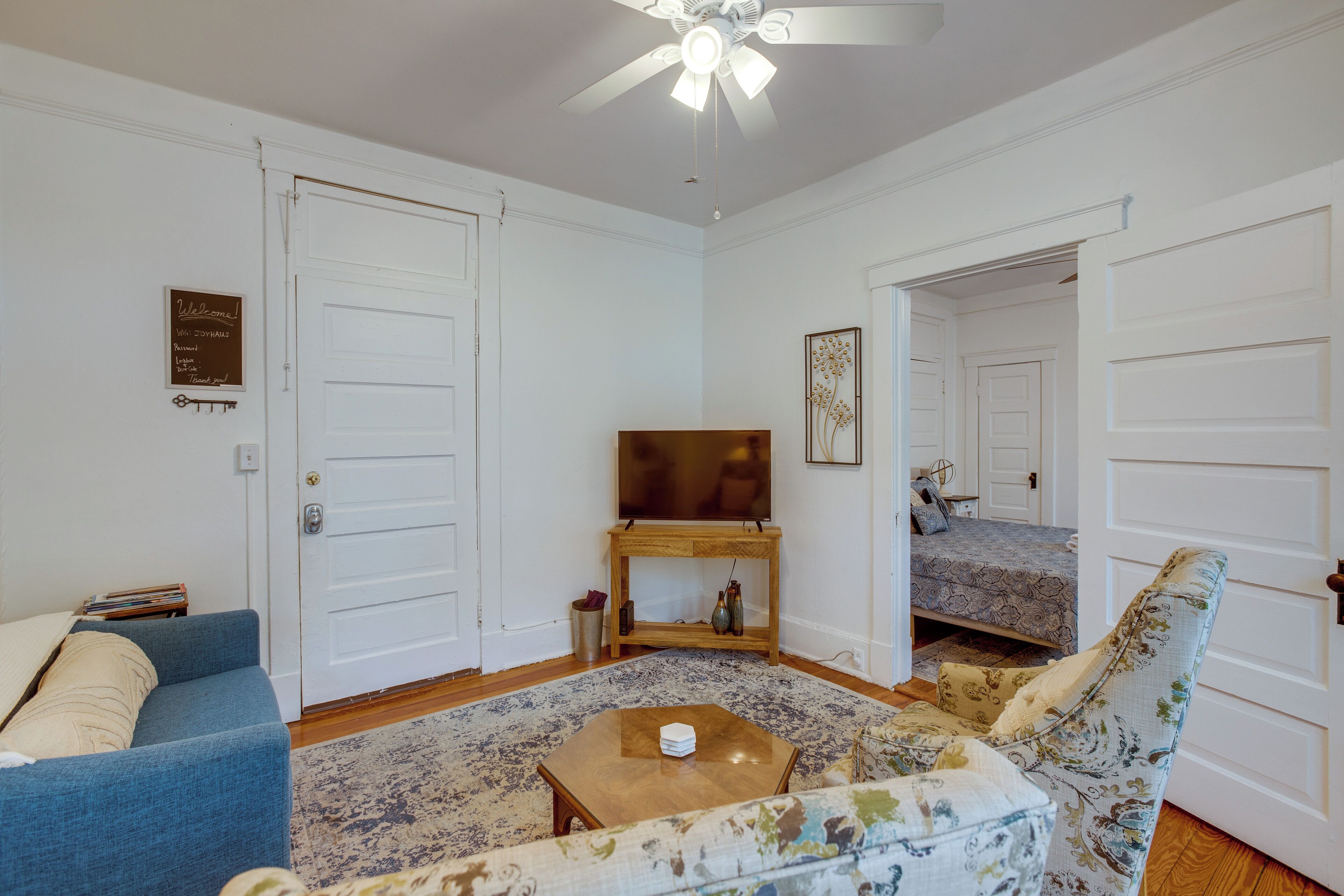 Un salon avec un canapé bleu, un fauteuil à motifs floraux, une table basse en bois et un téléviseur posé sur un meuble. Les portes sont blanches et il y a un ventilateur de plafond.
