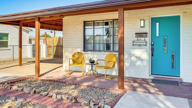 A covered patio with two yellow chairs and a small table, a turquoise door, and a brick pathway leading to the entrance. - Spacious Yard + Patio: Central Tucson Retreat! (Tucson)