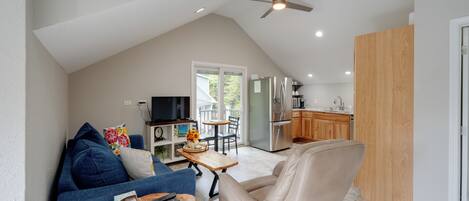 A modern loft-style living space with a high ceiling, a ceiling fan, a flat-screen TV, a dining area, and a kitchenette.