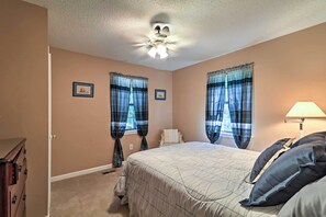 Une chambre à coucher comprenant un lit, un ventilateur de plafond, deux fenêtres avec des rideaux, une table de chevet et une lampe.