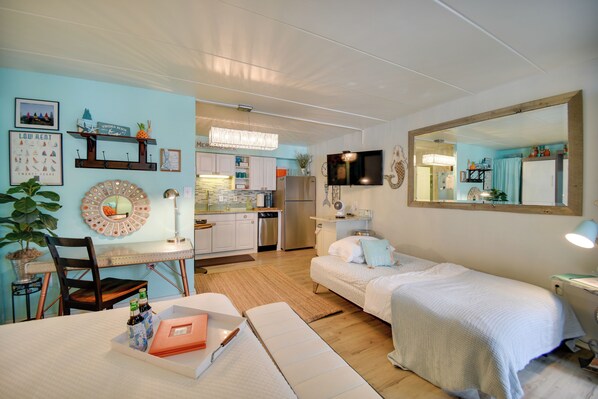 A cozy, well-lit room with a bed, bedside table, and a desk with a chair. A kitchen area is visible in the background.