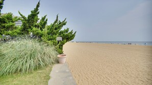 A sandy beach with a concrete path leading to a building. - Chic Virginia Beach Retreat - Pool, Steps to Beach (Virginia Beach)