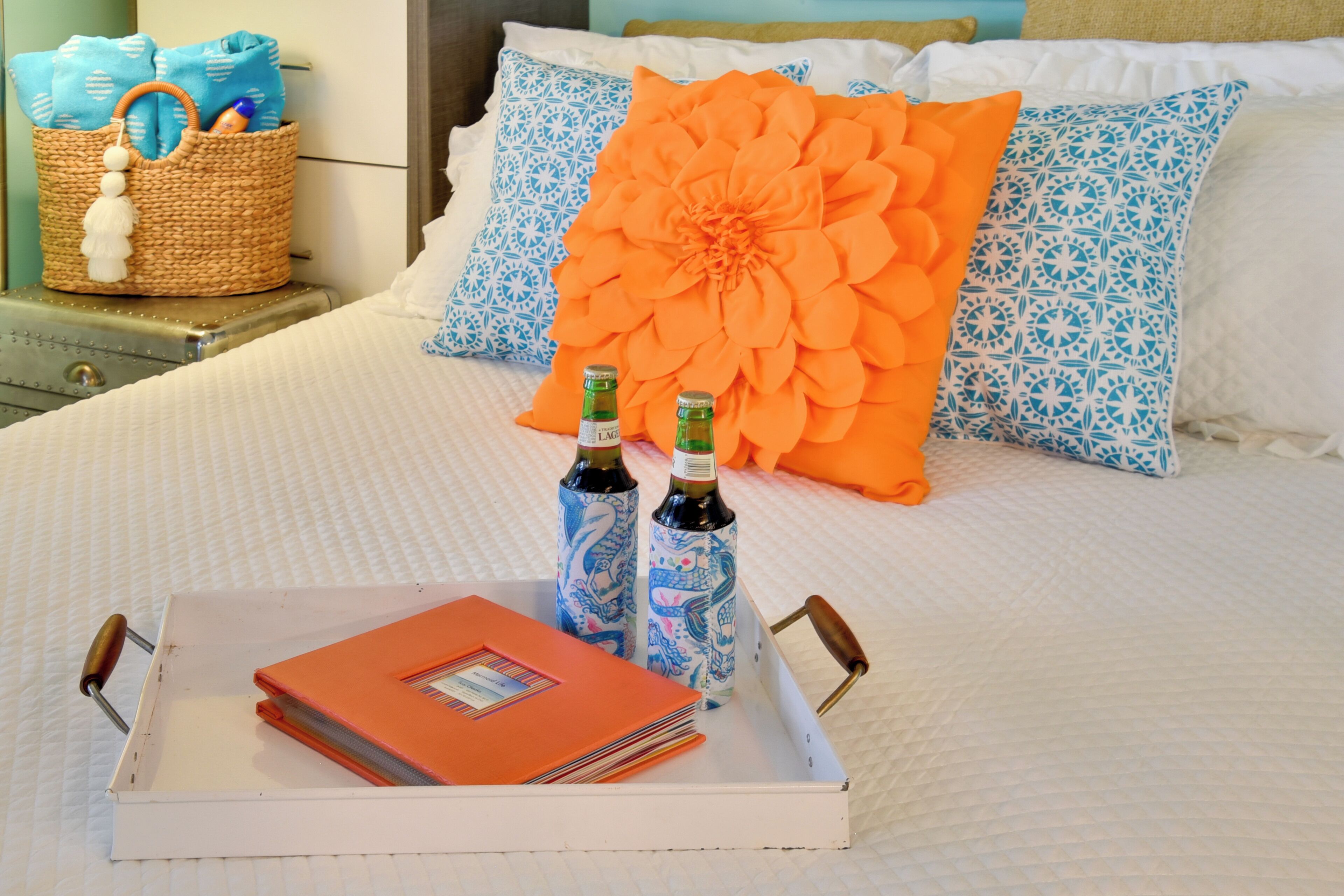 A neatly made bed with a large orange decorative pillow, two bottles of beer, and a tray with a red folder and a wicker basket.