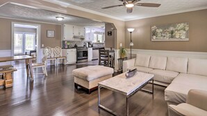 Amplia sala de estar con sofá, reposapiés y mesa de centro, un comedor con sillas y una cocina al fondo.
