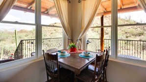 A dining area with a wooden table and chairs, a potted plant, and a view of a desert landscape through large windows with curtains. - Mountain Views & Private Jacuzzi: Tucson Casita (Tucson)