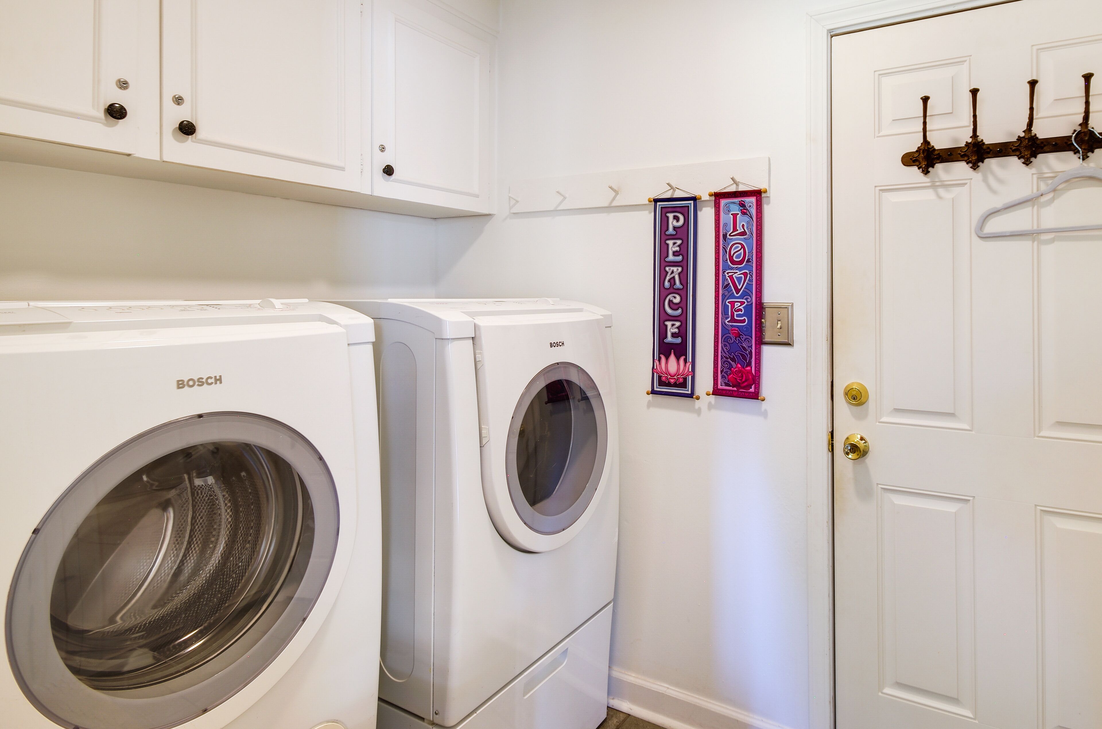 A laundry room with a Bosch washing machine and dryer, a white door with a coat rack, and two decorative signs on the wall.