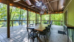 A covered outdoor dining area with a wooden table and chairs, surrounded by large windows offering a view of a forested area. - Stunning Culloden Cabin With Deck & Creek View! (Roberta)