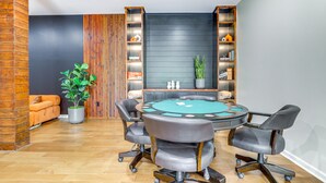 Una habitación con una mesa de póker, sillas, un sofá, una planta en maceta y estanterías con diversos objetos.