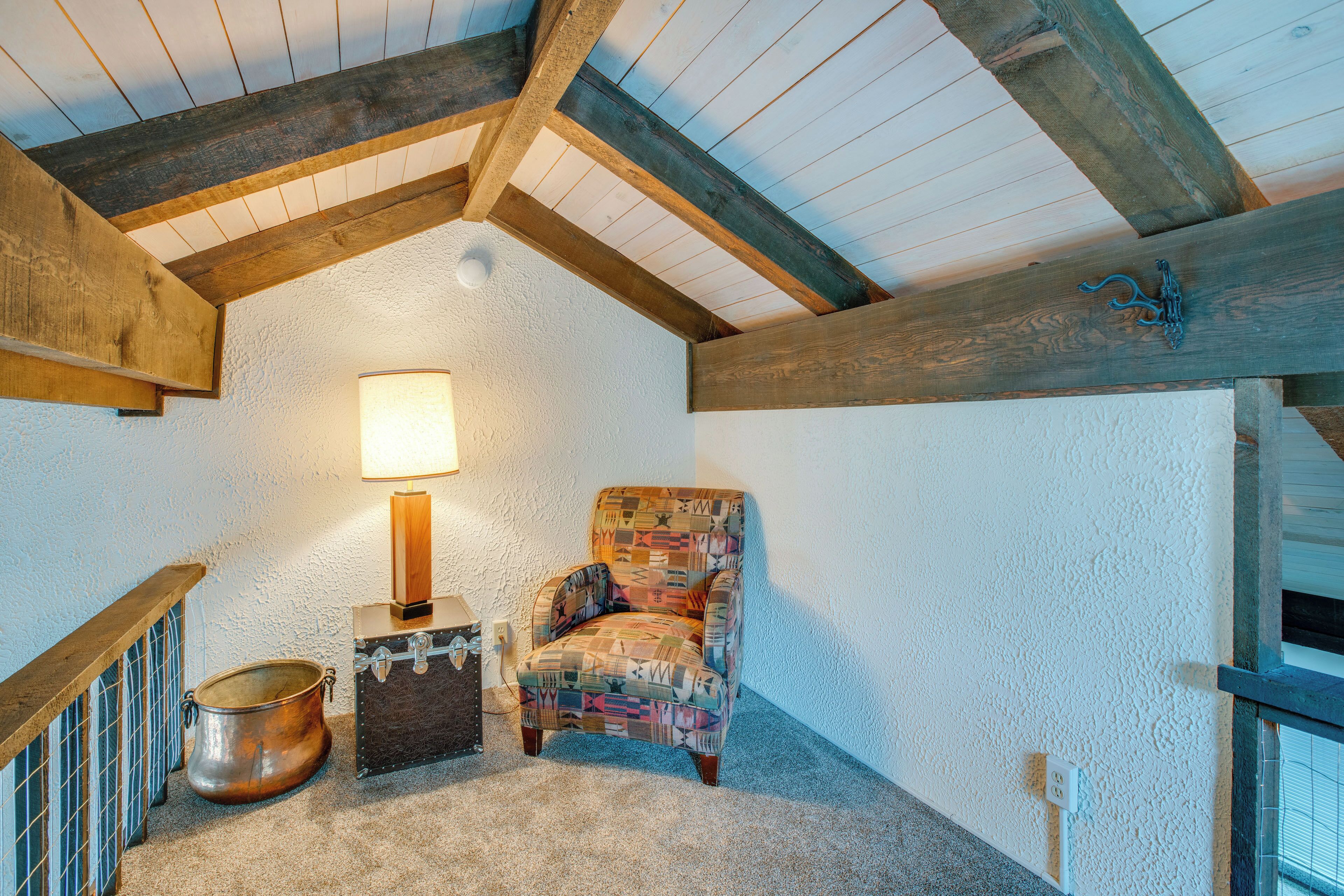 A cozy room with a high ceiling, exposed wooden beams, a cushioned armchair, a small table with a lamp, and a vintage suitcase.