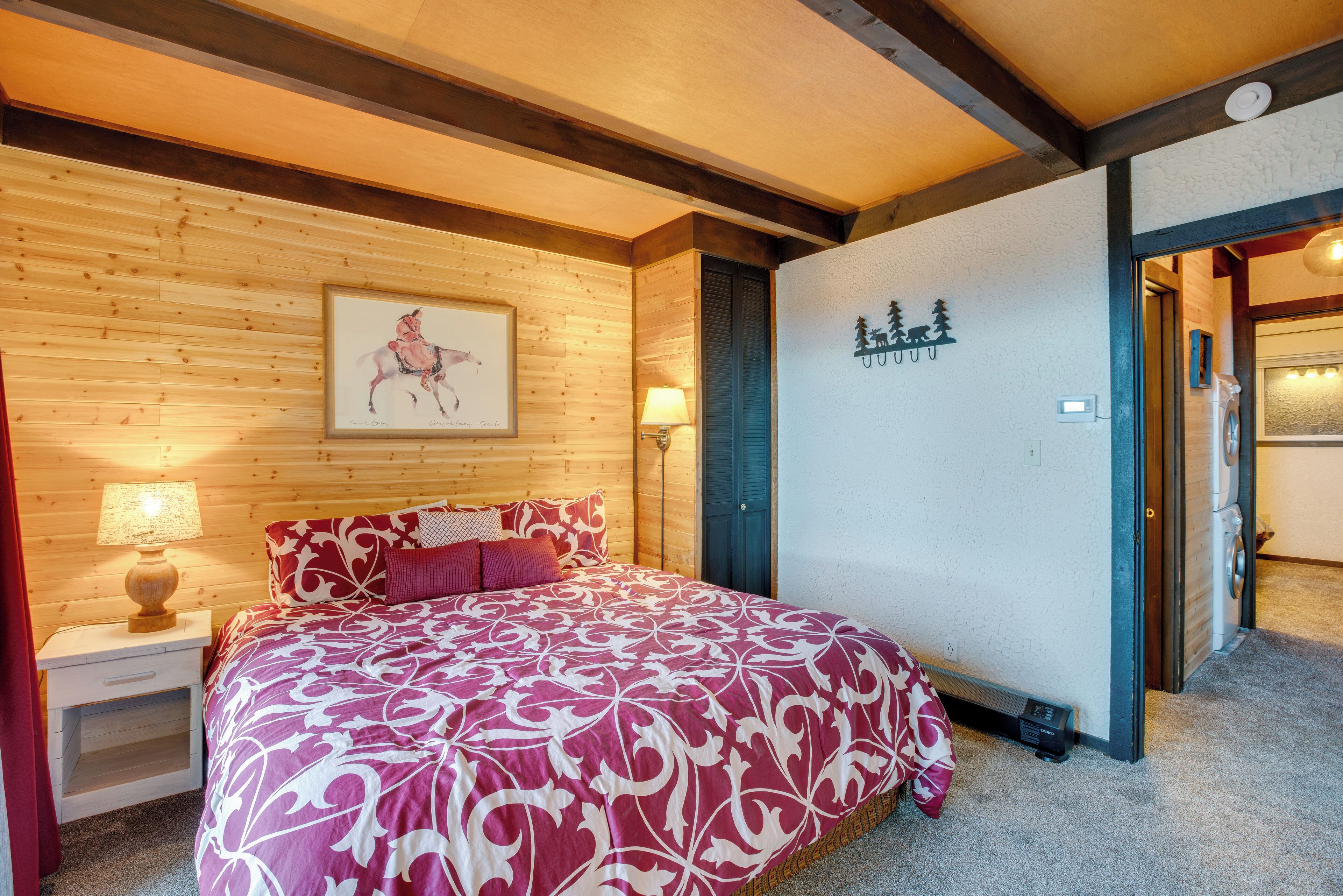 A bedroom with a wooden ceiling, a bed with a patterned bedspread, a nightstand with a lamp, and a wall-mounted artwork.