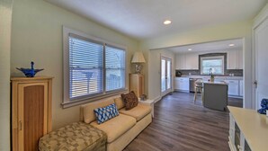A spacious living area with a sofa, ottoman, and a wooden cabinet. A kitchen with white cabinets and a central island is visible in the background. - Portsmouth Waterfront Vacation Rental w/ Deck (Portsmouth)