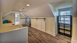 A loft-style room with a sloped ceiling, wooden flooring, a built-in bench with cushions, a window with blinds, and a door with a black metal handle. - Portsmouth Waterfront Vacation Rental w/ Deck (Portsmouth)