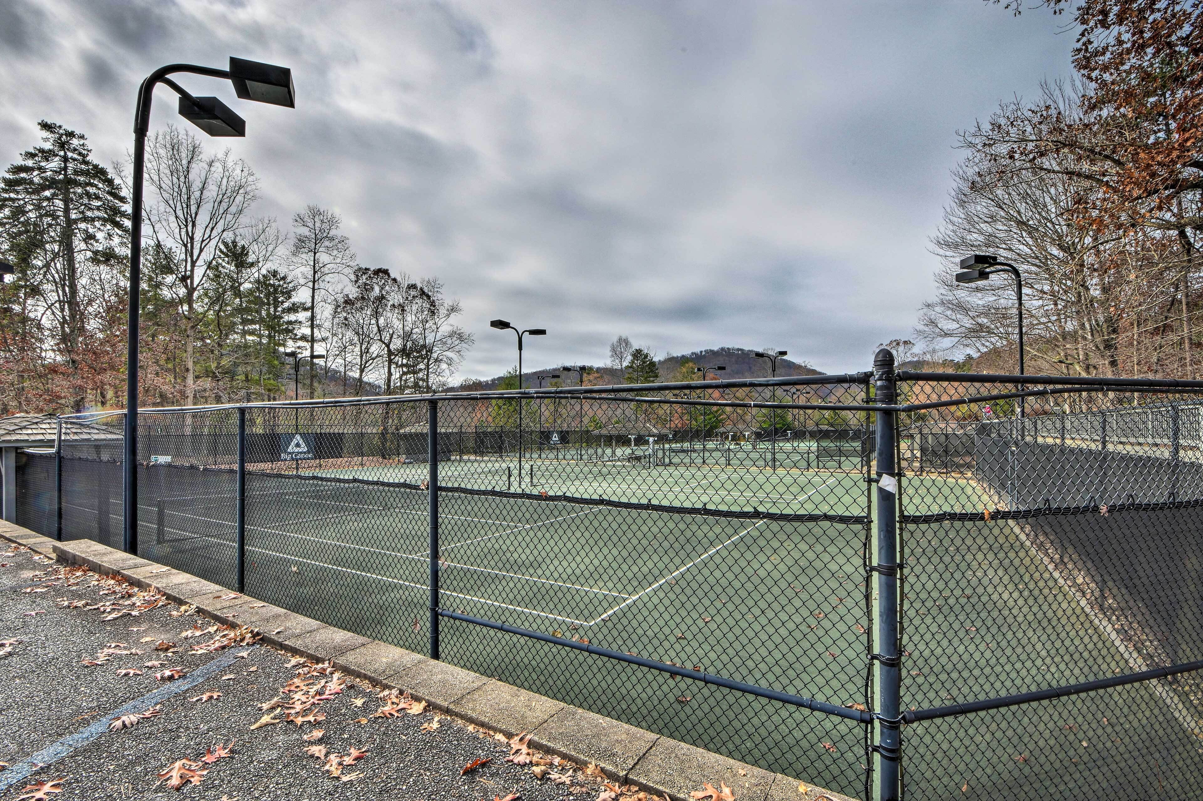 Una cancha de tenis con una cerca de malla, farolas y árboles al fondo.