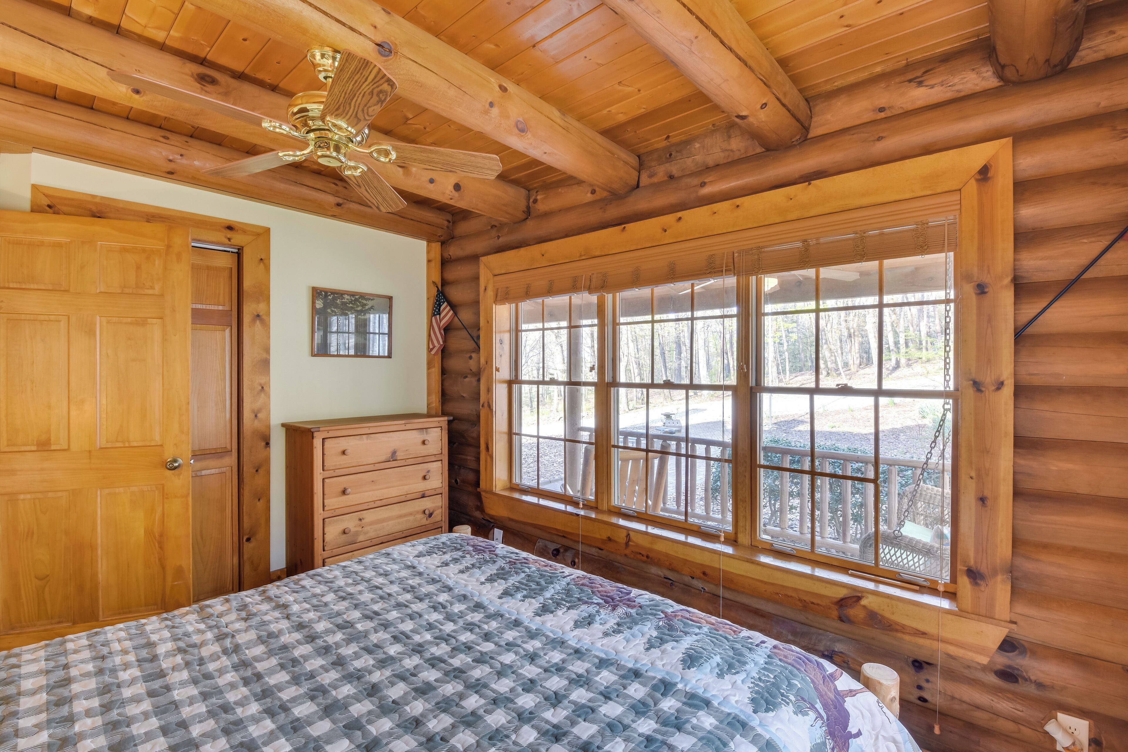 A log cabin bedroom with a bed, wooden dresser, and a large window with a view of the outdoors.