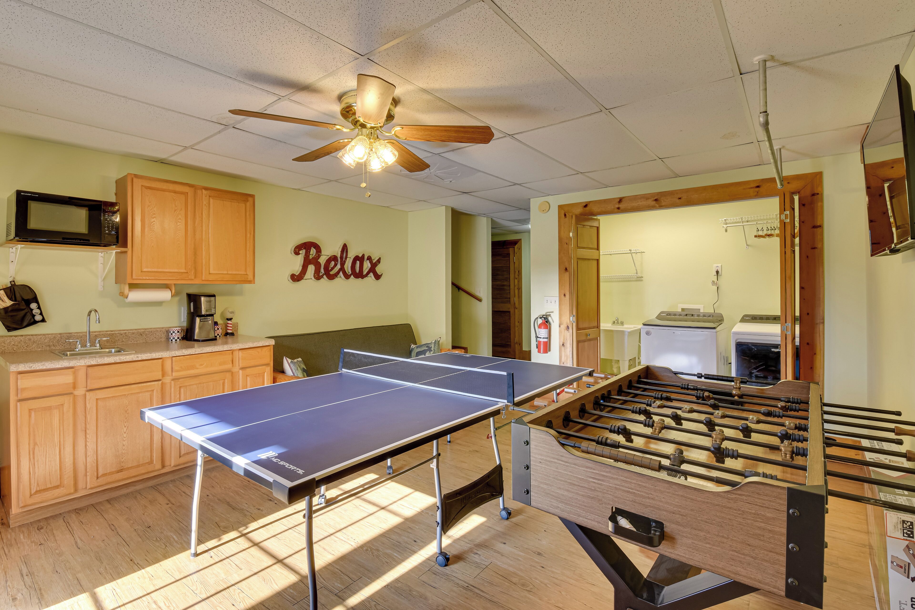 A room with a ping pong table, foosball table, wooden cabinets, a ceiling fan, and a microwave.