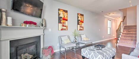Un salon avec une cheminée, un téléviseur à écran plat, deux fauteuils, un pouf à motifs et un escalier en arrière-plan.