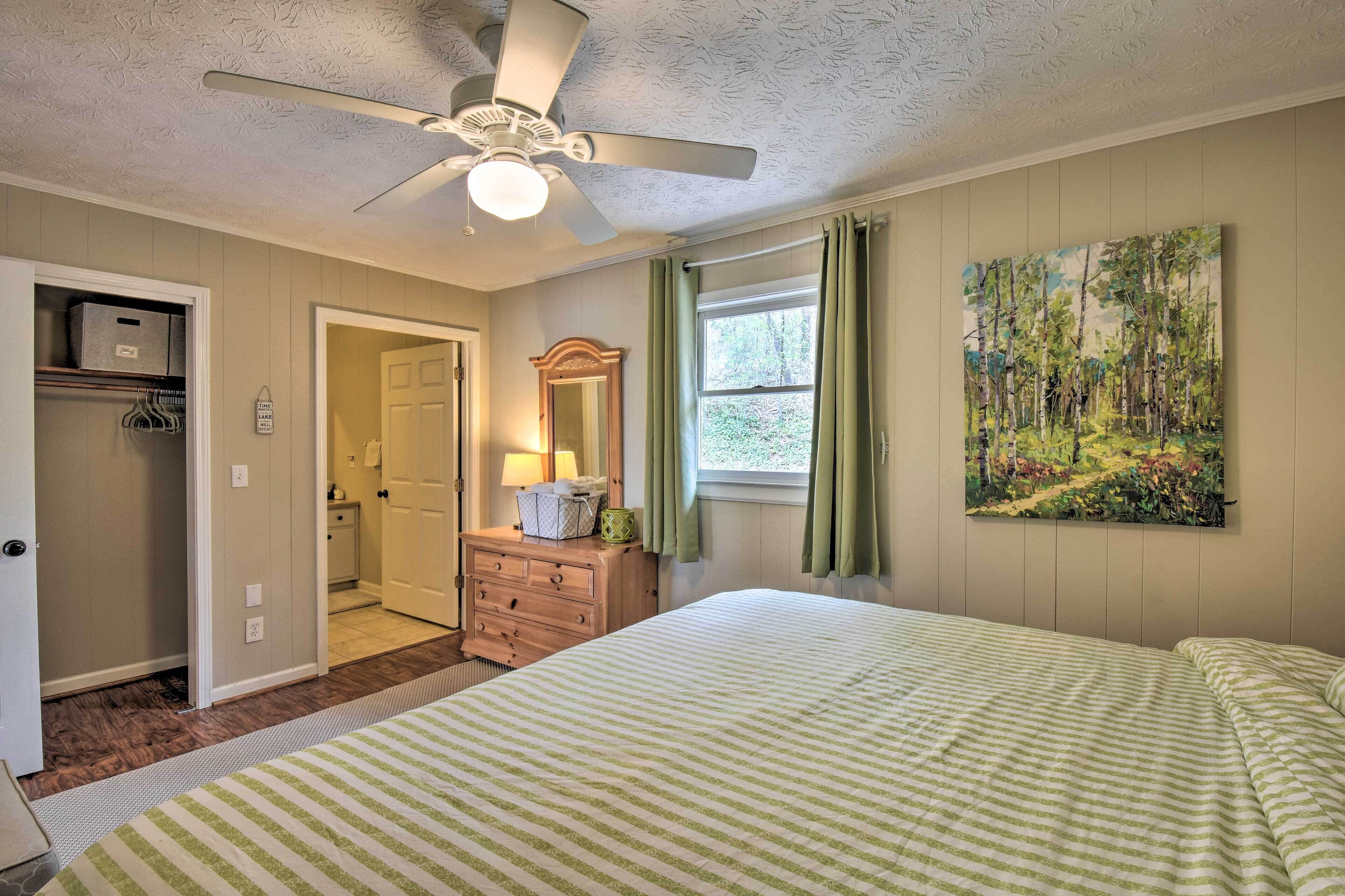 Une chambre à coucher comprenant un lit, une commode, un ventilateur de plafond, une fenêtre avec des rideaux et un tableau accroché au mur.