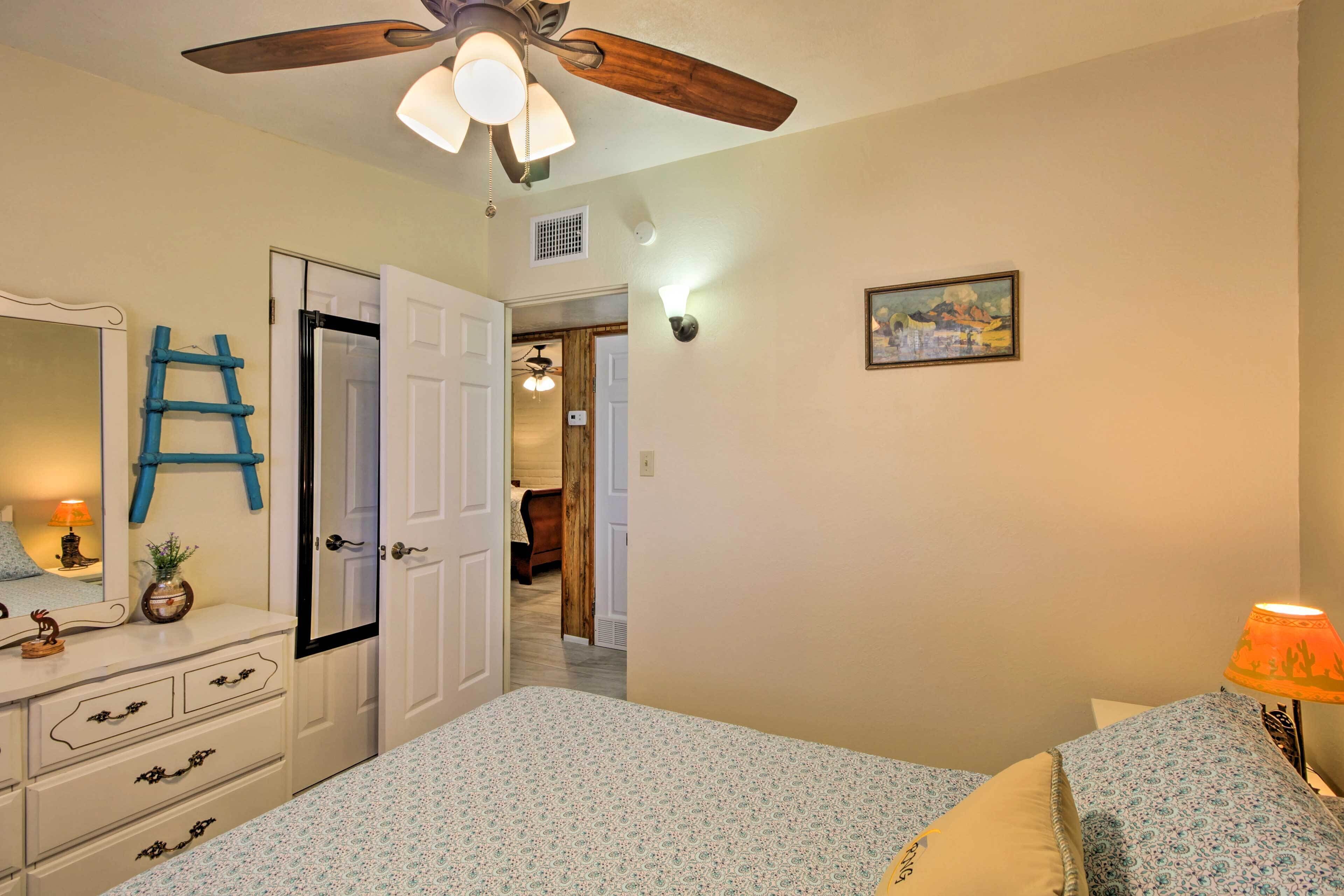 A bedroom with a bed, dresser, mirror, ceiling fan, and a hallway leading to another room.