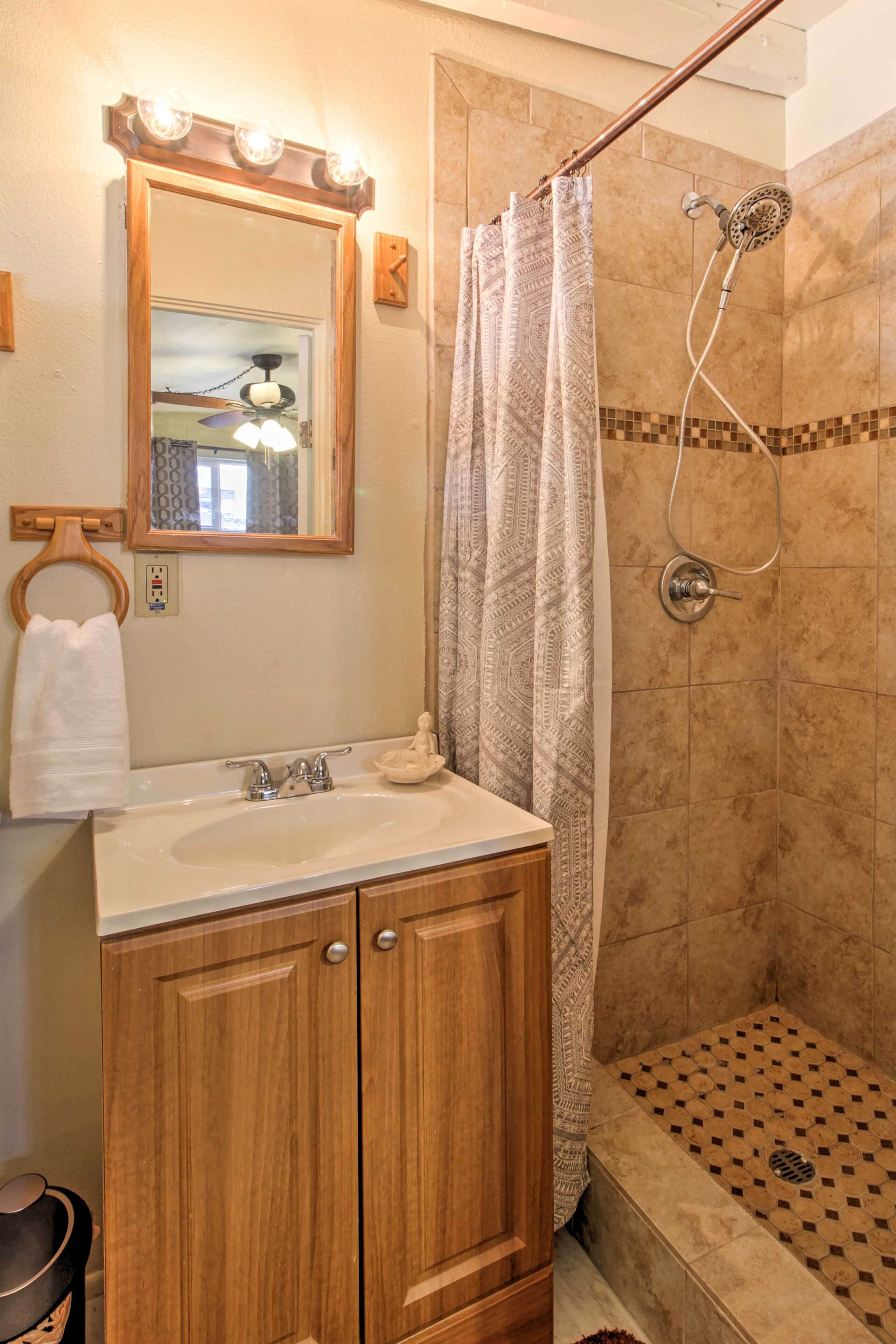 Une salle de bain avec un meuble-lavabo en bois, un évier, un miroir avec éclairage, une douche avec un rideau à motifs et un sol de douche carrelé.
