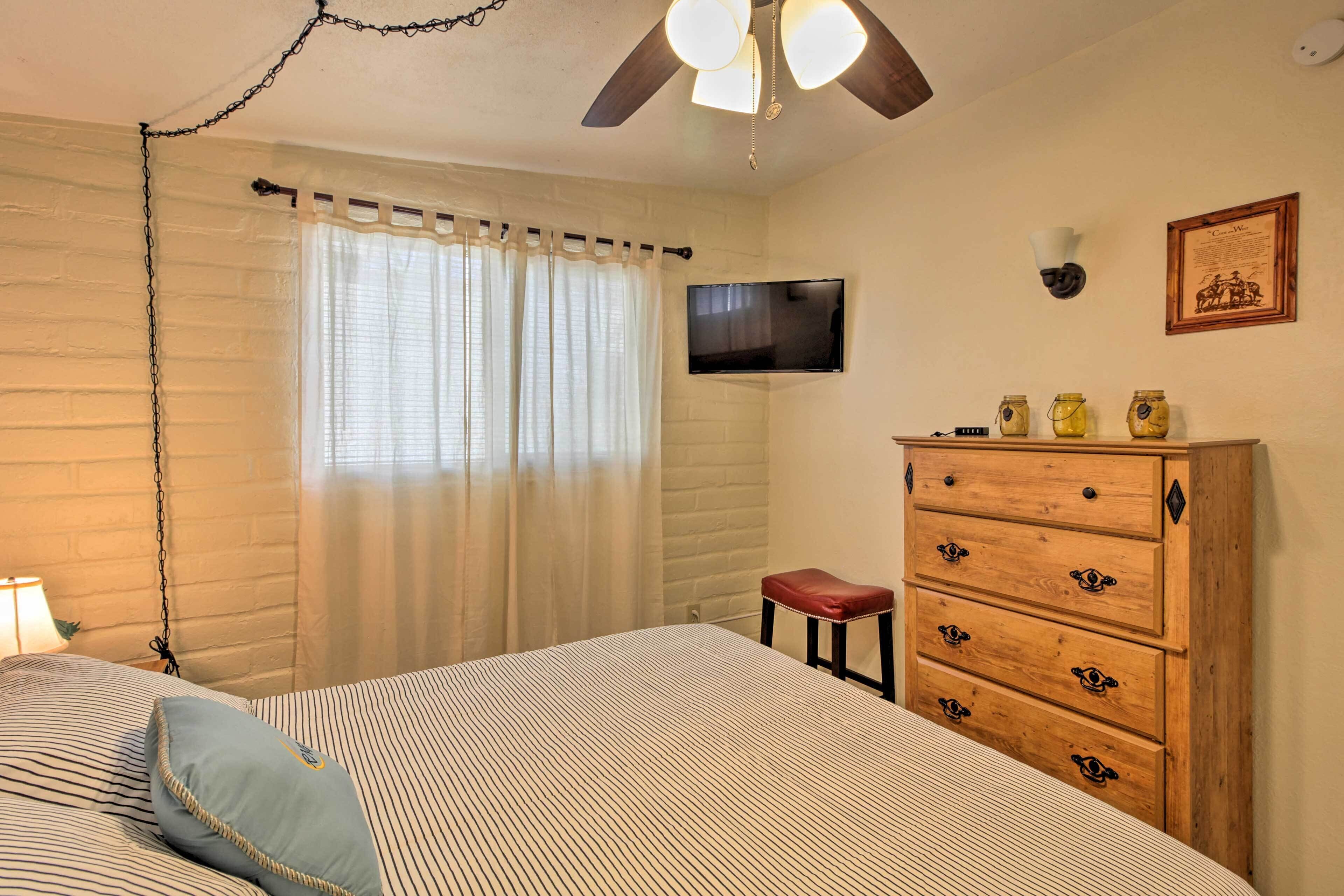 Une chambre à coucher avec un lit, une commode en bois, un téléviseur fixé au mur et une fenêtre avec des rideaux.