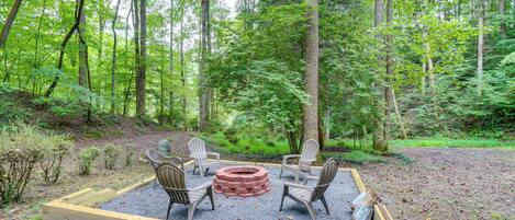 A circular fire pit surrounded by four Adirondack chairs in a forest setting.