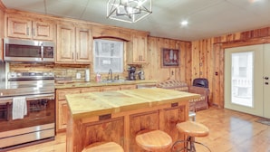 A kitchen with wooden cabinets, a large island with stools, stainless steel appliances, and a hanging light fixture. - Wooded Blue Ridge Cabin: 2 Decks, Fire Pit! (Cherry Log)
